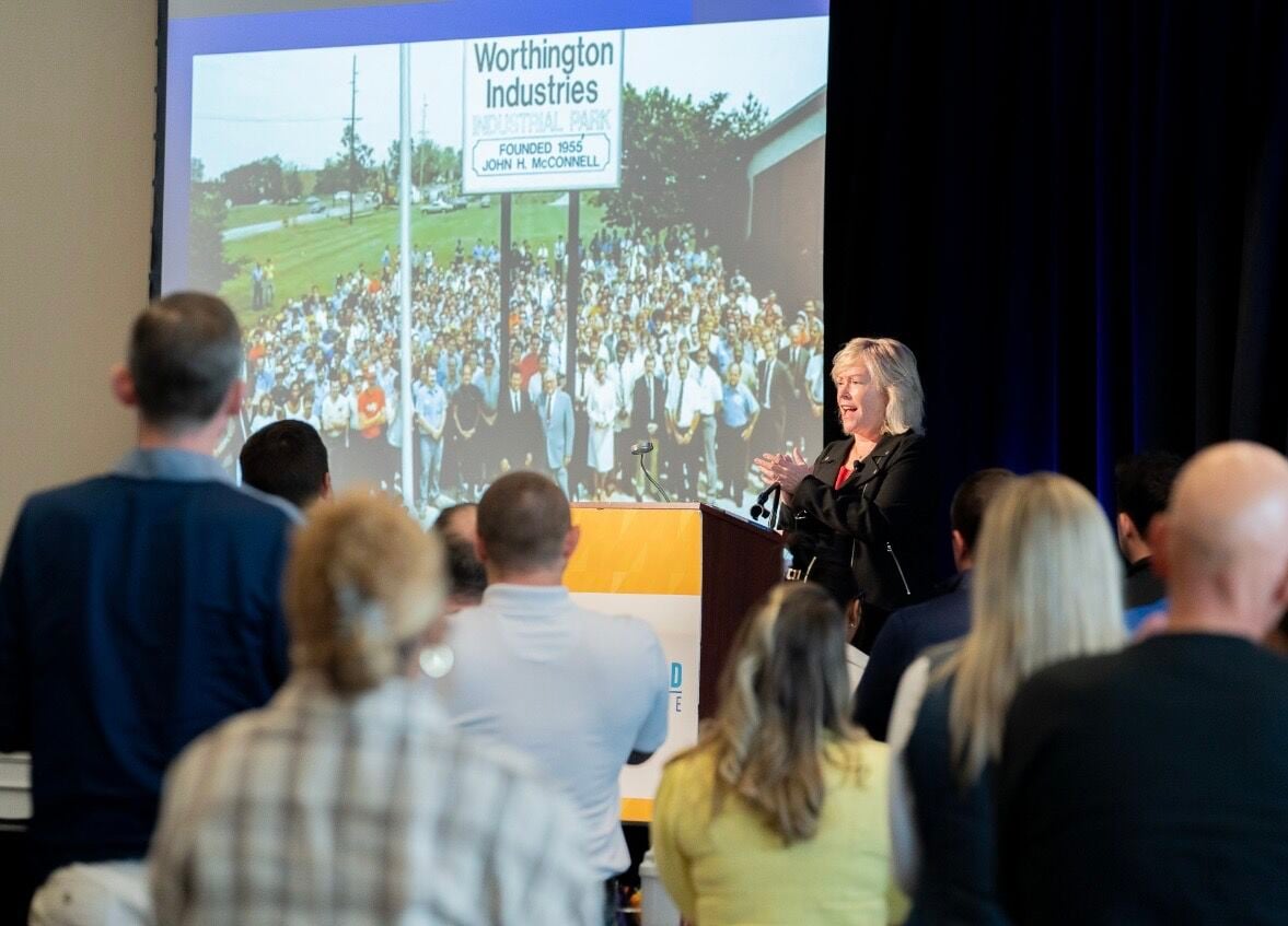 Woman speaking at podium on stage in front of crowd and projected screen