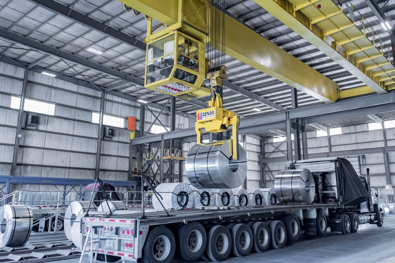 A master coil of galvanized steel being lowered from a yellow crane onto a flatbed truck.