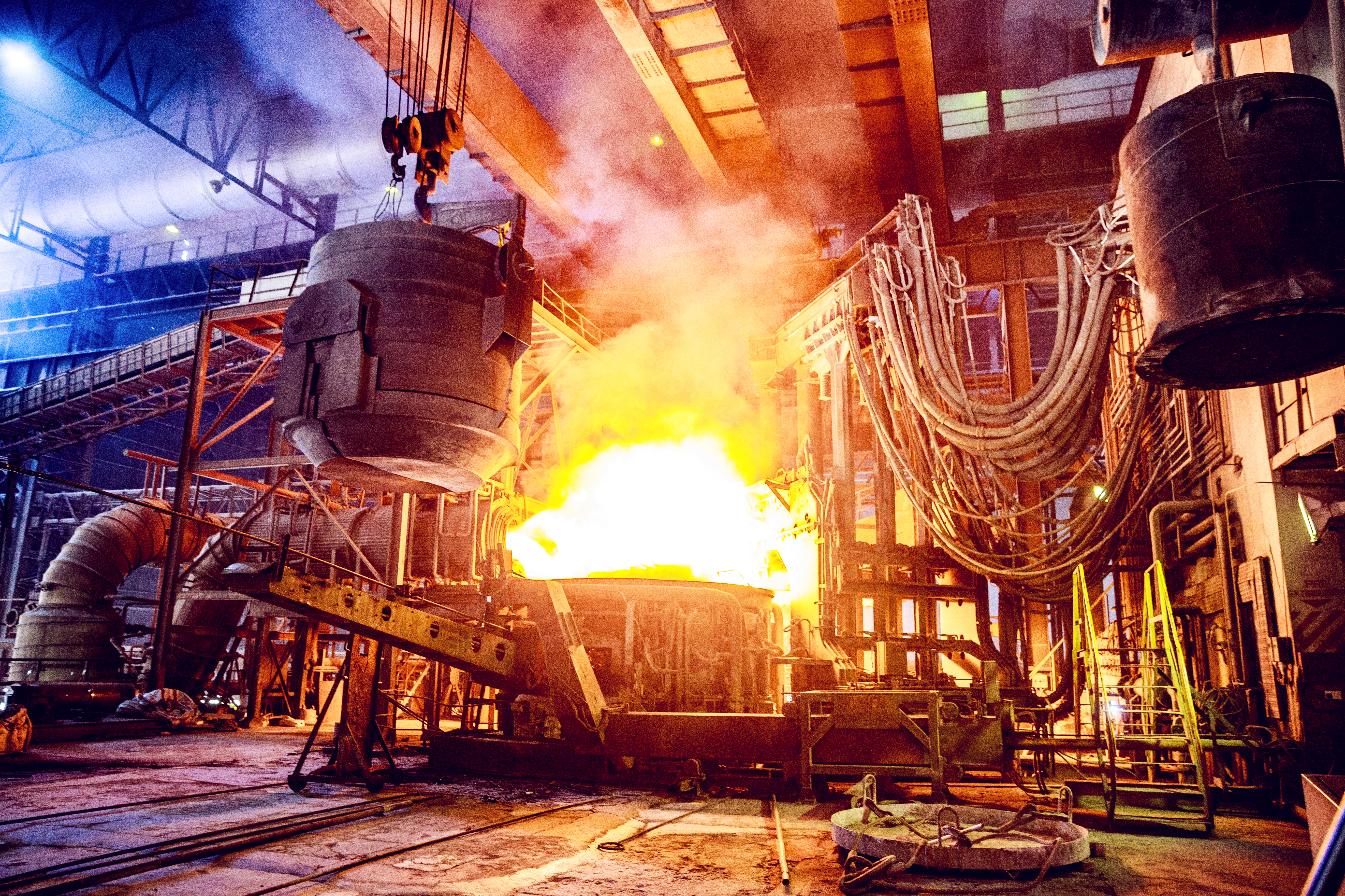 A large electric arc furnace sits at the middle of a facility floor with glowing orange molten steel in it.