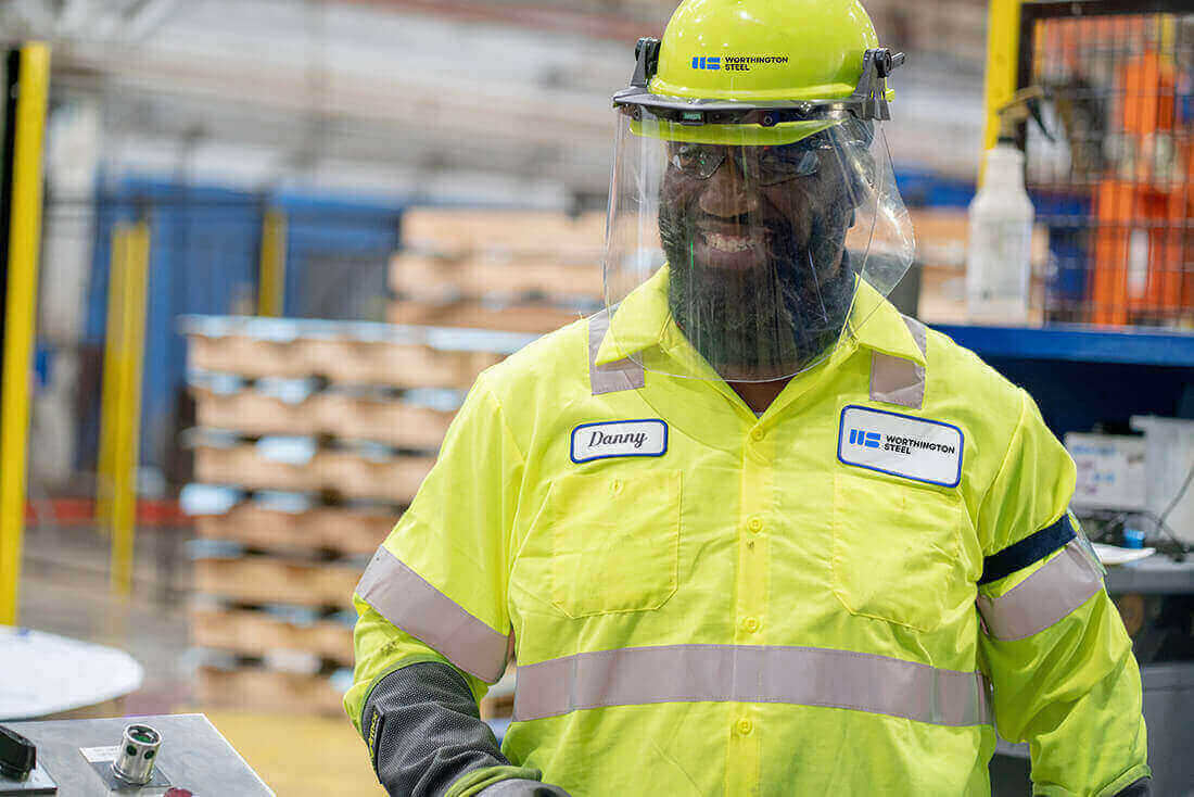 Worthington Steel employee wearing high-visibility safety gear and face shield inside a steel processing facility.